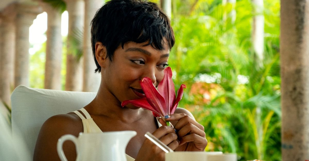 A woman sitting at a table sniffing a red flower.