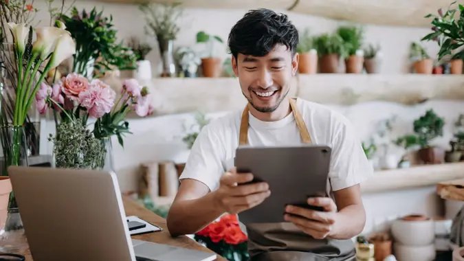 Asian male florist, small business flower shop owner, using digital tablet while working on laptop against flowers and plants.