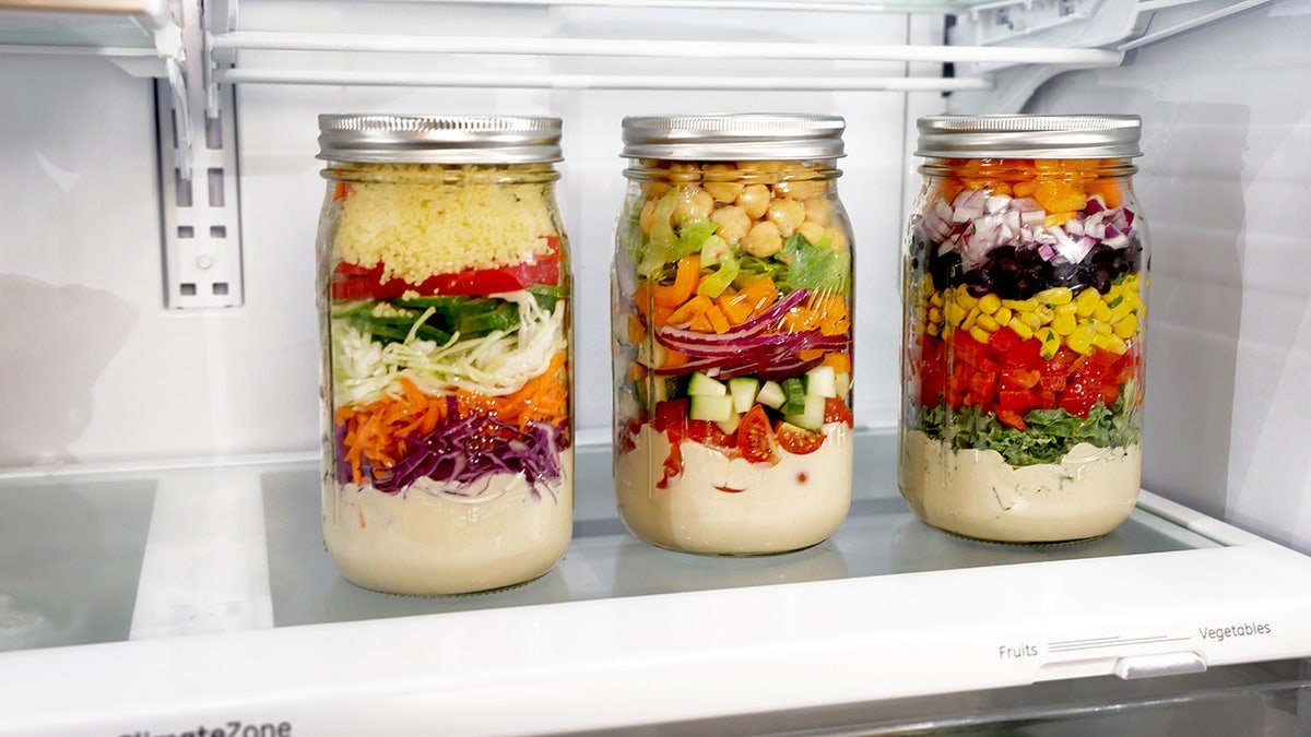 Three mason jars filled with "salads" which is layered with a dressing at the bottom. The individual layers are different in each jar, but contain vegetables, grains and other toppings.