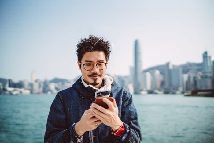 A man looks at his phone while standing along a waterfront with a city skyline behind him.